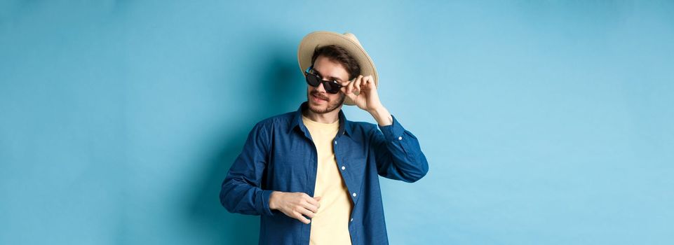 Handsome tourist in sunglasses and straw hat looking aside, smiling pleased, exploring sighsteeing on vacation, blue background.