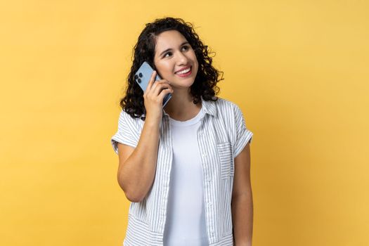 Portrait of smiling satisfied young adult beautiful woman with dark wavy hair standing and talking on smartphone, hearing good news. Indoor studio shot isolated on yellow background.