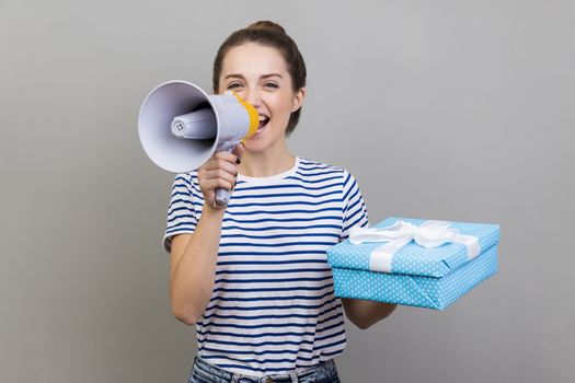 Portrait of pretty positive woman wearing striped T-shirt holding blue present box and making annoucement in megaphone, discounts. Indoor studio shot isolated on gray background.