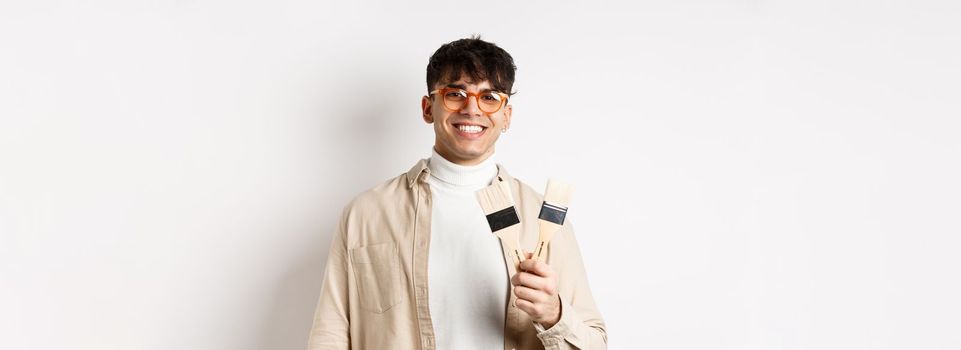 Hobbies and leisure concept. Stylish young man in glasses showing painting brushes and smiling, inviting to paint something, standing on white background.
