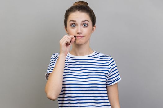 I won't tell anyone. Portrait of intimidated woman wearing striped T-shirt zipping lips and looking at camera, covering mouth promising to keep secret. Indoor studio shot isolated on gray background.
