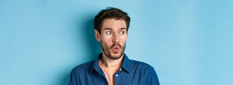 Close up of impressed man say wow and checking out banner, looking left at empty space amazed, standing on blue background.