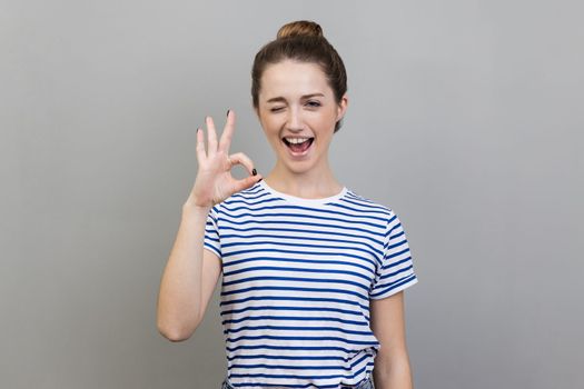 Portrait of funny playful positive young adult woman wearing striped T-shirt winking to camera and showing ok sign, approval gesture. Indoor studio shot isolated on gray background.