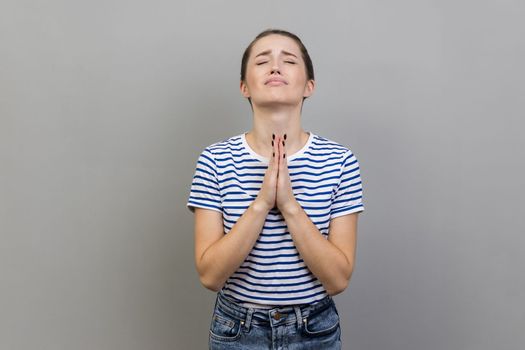 Please, I'm begging. Portrait of upset worried woman wearing striped T-shirt looking up with imploring desperate grimace, praying to god asking for help. Indoor studio shot isolated on gray background