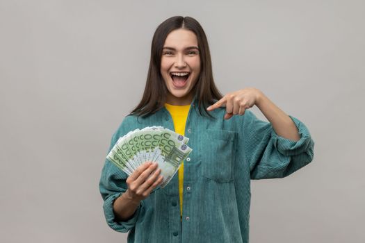 Delighted woman being happy to hold big money, pointing to euro bills in hand and looking with positive excited expression, wearing casual style jacket. Indoor studio shot isolated on gray background.