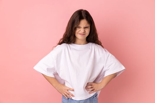 Angry aggressive little girl wearing white T-shirt standing with hands on hips, looking at camera with frowning face, expressing anger and hate. Indoor studio shot isolated on pink background.