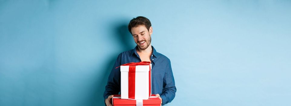 Romantic young man with beard, looking happy at gift boxes on valentines day, giving presents to lover, standing over blue background.