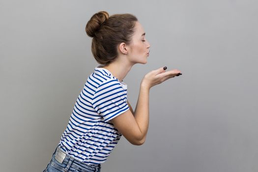 Side view of charming amorous woman in striped T-shirt giving air kiss to camera over opened palms, kissing with pout lips, sharing romance and love. Indoor studio shot isolated on gray background.