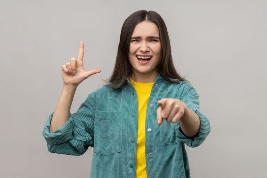 Disrespectful rude young woman showing loser gesture and pointing on you, abuser, mocking your failures, wearing casual style jacket. Indoor studio shot isolated on gray background.