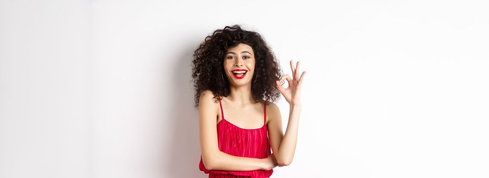 Cheerful caucasian woman with curly hair and makeup, wearing elegant red dress, showing OK sign and smiling in approval, standing over white background.