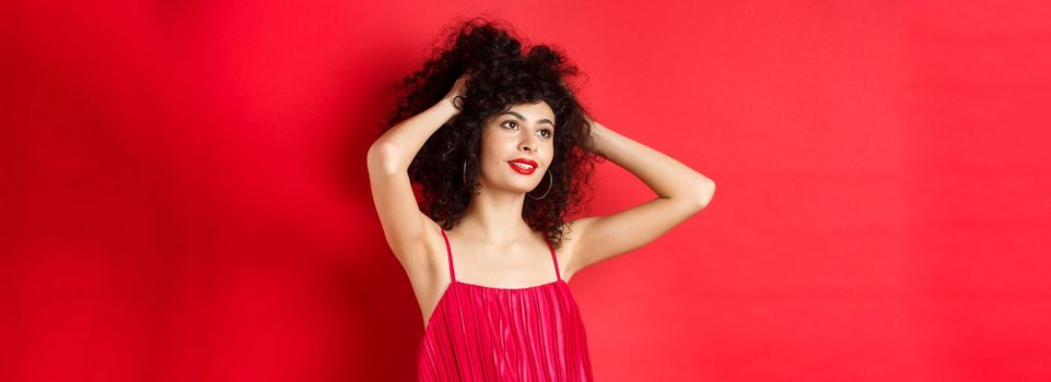 Carefree female model playing with hair, looking dreamy and relaxed at logo, smiling sensual, standing on red background.