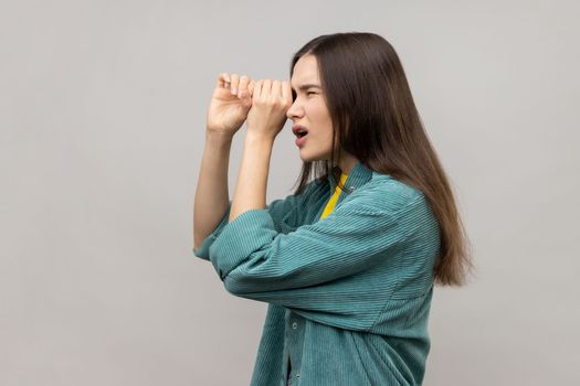 Side view of shocked young woman standing with hands on eye monocular gesture and looking away with surprised face, wearing casual style jacket. Indoor studio shot isolated on gray background.