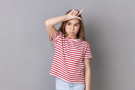 Portrait of depressed little girl wearing striped T-shirt showing loser gesture, L finger sign on forehead, upset about dismissal, unlucky day. Indoor studio shot isolated on gray background.