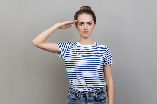 Yes, sir. Portrait of woman wearing striped T-shirt saluting with respect and responsibility, patriotic citizen listening order with honor. Indoor studio shot isolated on gray background.