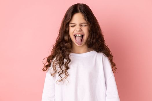 Portrait of childish carefree little girl wearing white T-shirt showing out tongue and closing eyes with naughty disobedient grimace, making face. Indoor studio shot isolated on pink background.