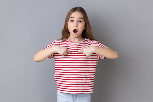 Portrait of astonished little girl wearing striped T-shirt pointing at herself, asks who me, has surprised expression, shocked being picked. Indoor studio shot isolated on gray background.