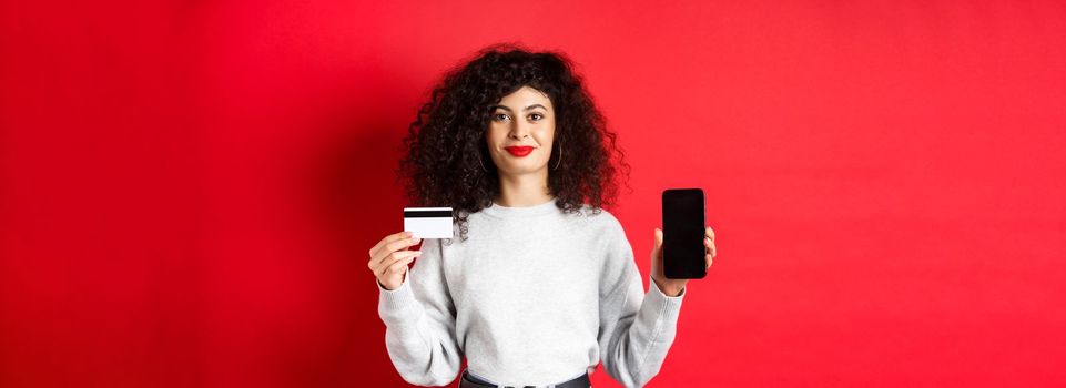 Young modern woman with curly hair showing plastic credit card and mobile phone screen, demonstrating online shopping app, standing on red background.