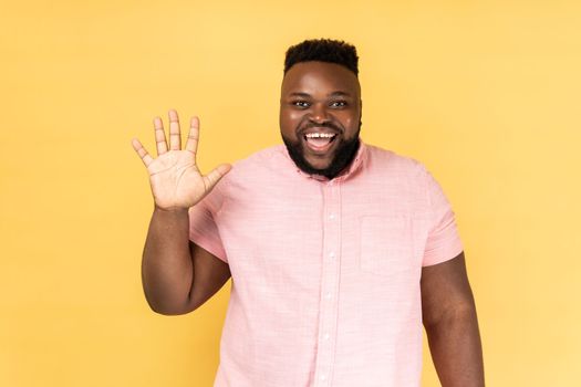 Portrait of friendly bearded man in pink shirt greeting you rising hand and waving, saying hi, glad to see you, looking at camera with toothy smile. Indoor studio shot isolated on yellow background.
