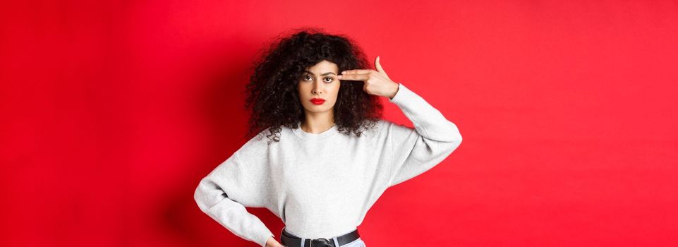 Annoyed caucasian woman with curly hair, showing hand gun sign on head, blow her mind off, standing bothered against red background.