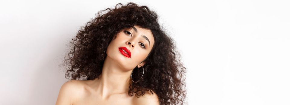 Close-up portrait of sensual and romantic woman with curly hair, red lips, looking seductive at camera, posing on white background.