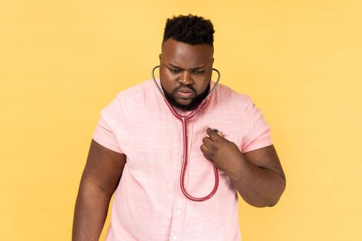 Portrait of concentrated bearded man wearing pink shirt standing with stethoscope, doing self examining of his heart, medicine concept. Indoor studio shot isolated on yellow background.
