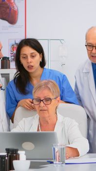 Medical team discussing diagnosis about patients treatment issues record in workplace using laptop. Group of doctors, teamwork people having health brainstorming conference in hospital meeting room.