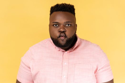Portrait of funny positive young adult man wearing pink shirt standing with pouted lips making fish face, having fun, childish good mood. Indoor studio shot isolated on yellow background.