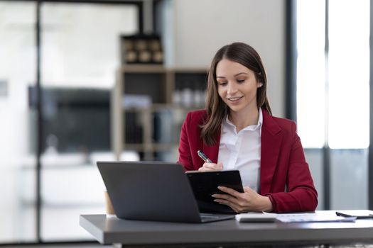 Smiling woman sitting at desk working on laptop taking notes in notebook, happy millennial female working online, watching webinar using computer and writing check list.