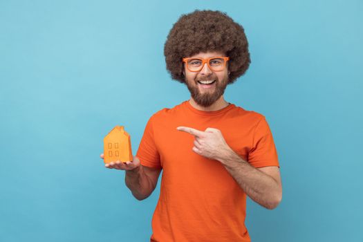 Portrait of smiling man with Afro hairstyle wearing orange T-shirt and funny eyeglasses holding pointing at paper house, helping with rent. Indoor studio shot isolated on blue background.