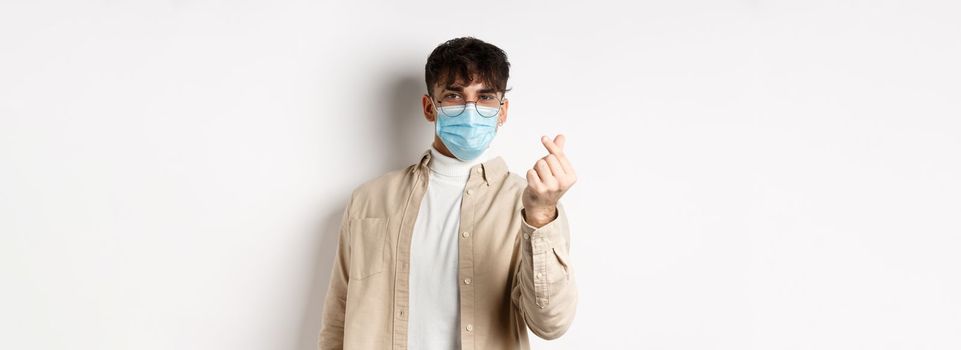 Covid-19, health and real people concept. Cute young man in medical mask showing finger heart and look at camera, standing on white background.