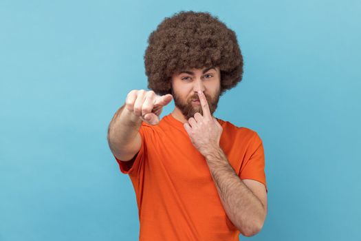 Portrait of man with Afro hairstyle wearing orange T-shirt touching nose, showing liar gesture, angry about falsehood, pointing to camera fake news. Indoor studio shot isolated on blue background.