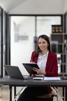 Portrait smiling woman sitting at desk working on laptop taking notes in notebook, happy millennial female working online, watching webinar using computer and writing check list.