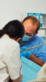 Stomatologist with face mask and gloves using drill tool for cleaning teeth of little patient working together with man assistent in modern stomatological clinc. Mother visiting dentist with child.