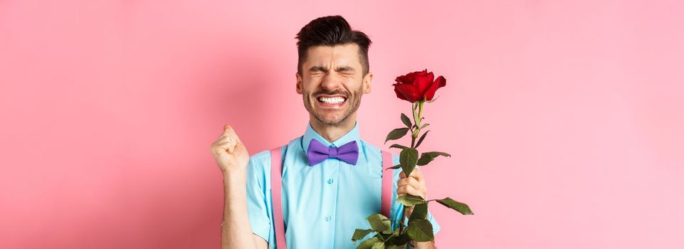 Romance and Valentines day concept. Hopeful man feeling excited before date, waiting for lover with red rose and praying, standing over pink background.