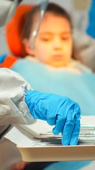 Medical team working with sterilized tools wearing face shield, coverall suit, mask and gloves during coronavirus outbreak in new normal stomatological clinic. Dentist treating teeth of little girl