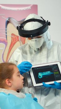 Dentist with ppe suit pointing on digital screen explaining x-ray to mother of girl patient. Medical team and patients wearing face shield coverall, mask, gloves, showing radiography using notebook