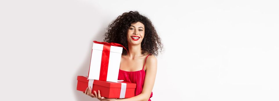 Beautiful birthday girl with curly hair, holding bday gifts and smiling happy, celebrating, standing against white background.