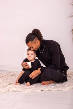 Stylish father and son, in black clothes, sitting on a knitted blanket, on the floor in the room. A father embraces his sons . Copy space. Vertical