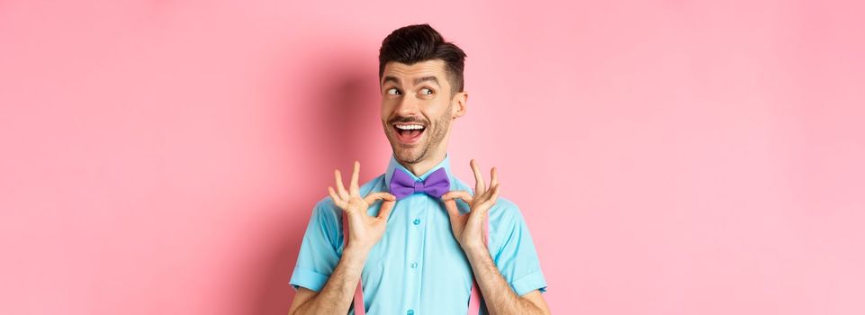 Cheerful and confident young man with moustache, fixing bow-tie on neck and looking left pensive, smiling happy, standing on pink background.
