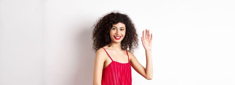 Beauty and fashion. Friendly elegant woman with curly hair, red dress, waving hand and saying hello, smiling to greet you, standing on white background.