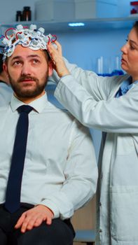 Woman researcher putting brainwave scanning headset to patient developing experiments using brain study lab monitors. Man siting on chair while scientist adjusting device examining nervous system