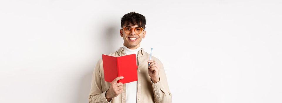 Happy person in glasses writing in journal, holding pen and diary, smiling at camera, plan a schedule, standing with planner on white background.