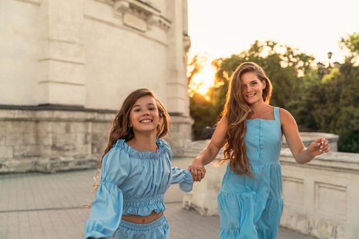 Portrait of a mother and daughter in blue dresses with flowing long hair against the backdrop of a sunset and a white building. They run and smile at the camera. Family stories on the weekend.