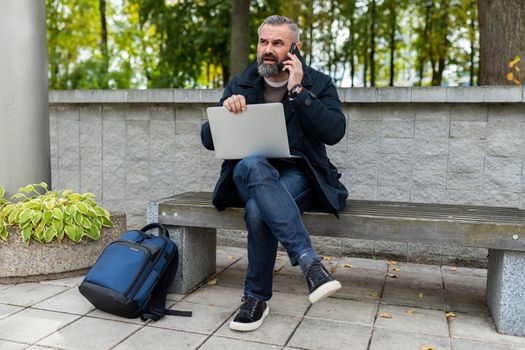 mature man working on laptop outside talking on the phone.