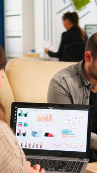 Close up of back view young entrepreneur sitting on couch in office using laptop computer while colleagues working on background. Multiethnic coworkers planning new financial project in modern company