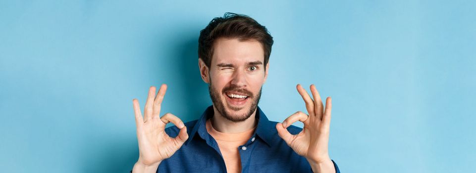 Close up of handsome confident guy winking, showing okay gestures, guarantee quality, recommending something good, standing on blue background.