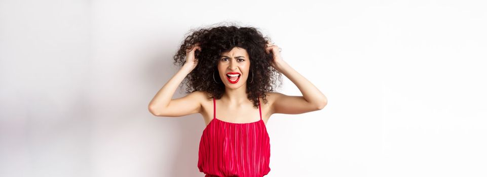 Frustrated curly woman in red dress, frowning and screaming angry, pull out hair and shout at camera, standing on white background.