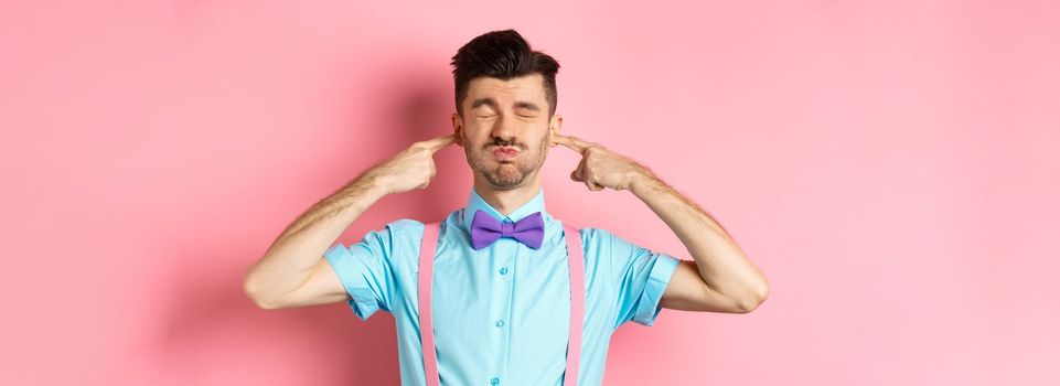 Annoyed funny guy in bow-tie shut ears, plug fingers and grimacing, irritated by loud noise, standing over pink background.