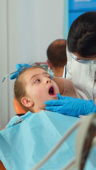Pediatric dentist treating teeth to little girl patient in clinic lying on stomatological chair with open mouth. Doctor and nurse working together in stomatological office wearing protection mask