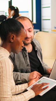 Diverse business people working in teamwork discussing project strategy using laptop sitting on couch at small table. Manager executive analysing financial data from black woman tablet at team meeting